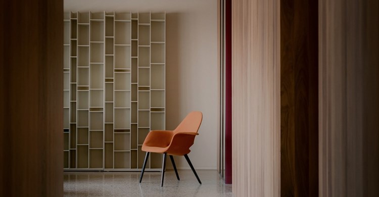 Una stanza in cui l'arredo moderno e minimalista evidenzia il vuoto della libreria bianca a vista, in contrasto con una sedia arancione posta a fianco